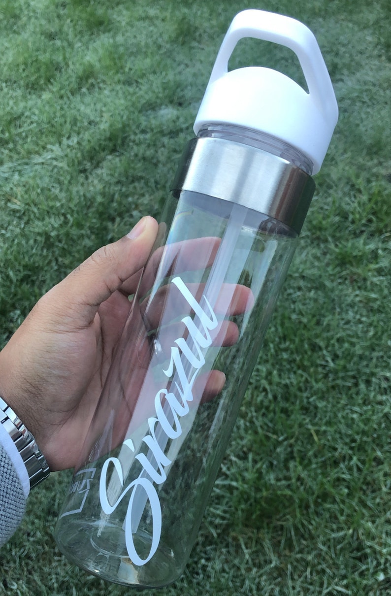 May include: A clear plastic water bottle with a white lid and a silver band around the middle. The bottle has a white straw and the word "Snazzy" written on the side in white cursive.
