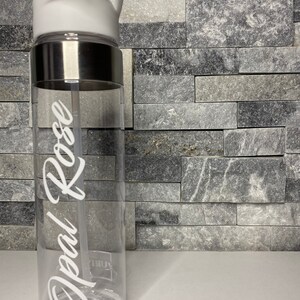 May include: A clear plastic water bottle with a white lid and a silver band around the middle. The bottle has a white straw and the name "Opal Rose" written on the side in white cursive.