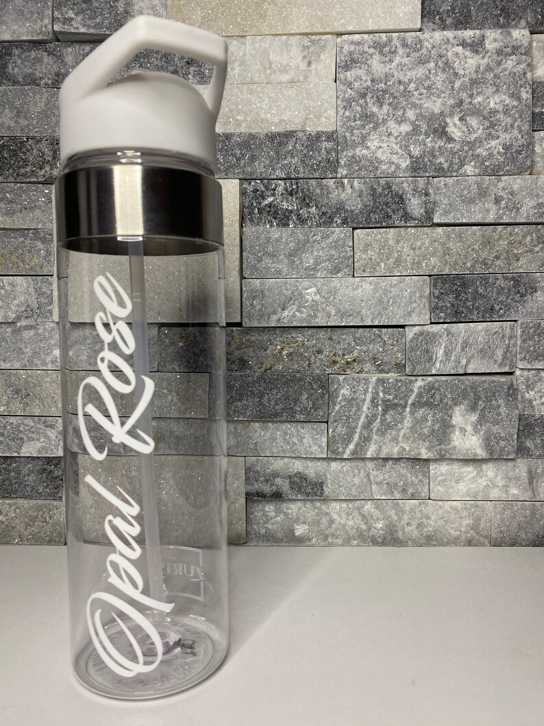 May include: A clear plastic water bottle with a white lid and a silver band around the middle. The bottle has a white straw and the name "Opal Rose" written on the side in white cursive.
