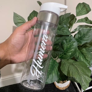 May include: Clear plastic water bottle with a silver lid and a white straw. The bottle has the name "Humera" written on it in white cursive.