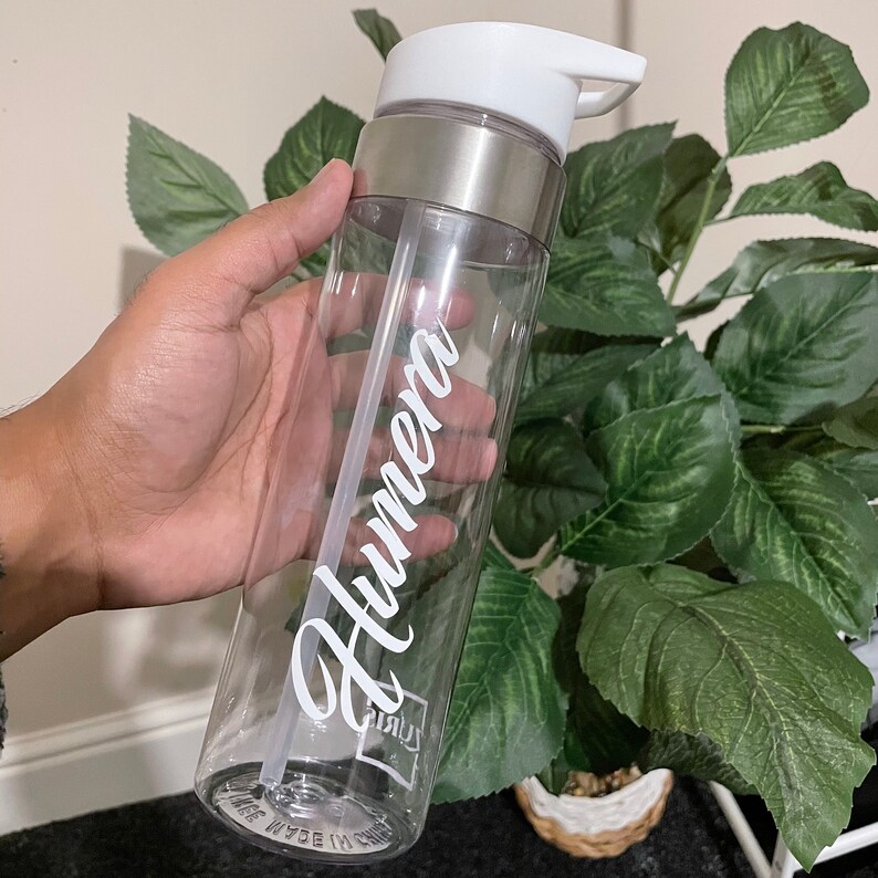 May include: Clear plastic water bottle with a silver lid and a white straw. The bottle has the name "Humera" written on it in white cursive.