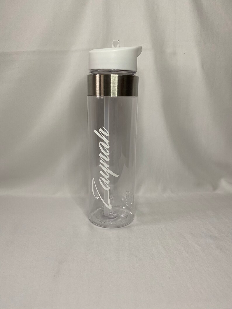 May include: A clear plastic water bottle with a silver band around the middle and a white lid. The bottle has a straw and the name "Laynah" is written on the side in white.