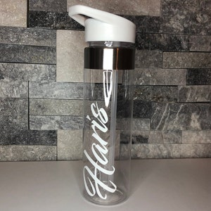 May include: A clear plastic water bottle with a white lid and a silver band around the middle. The bottle has the name "Haris" written on it in white.