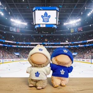 May include: Two plush figures wearing Toronto Maple Leafs apparel, including hats and sweaters with the team logo. They stand on a wooden surface in front of a hockey arena with a large scoreboard and cheering fans.