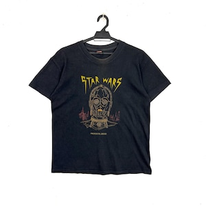 Puede incluir: Camiseta negra de Star Wars con una ilustración dorada y roja de C-3PO y la palabra "STAR WARS" en amarillo. El texto "PROTOCOL DROID" está debajo. La camiseta está colgada de una percha negra.