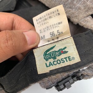 Puede incluir: Un gorro Lacoste gris y negro con un logotipo de cocodrilo verde. El gorro tiene una etiqueta de talla que dice "M 56,5 cm" y "日本製", que se traduce como "Hecho en Jap&oacute;n".
