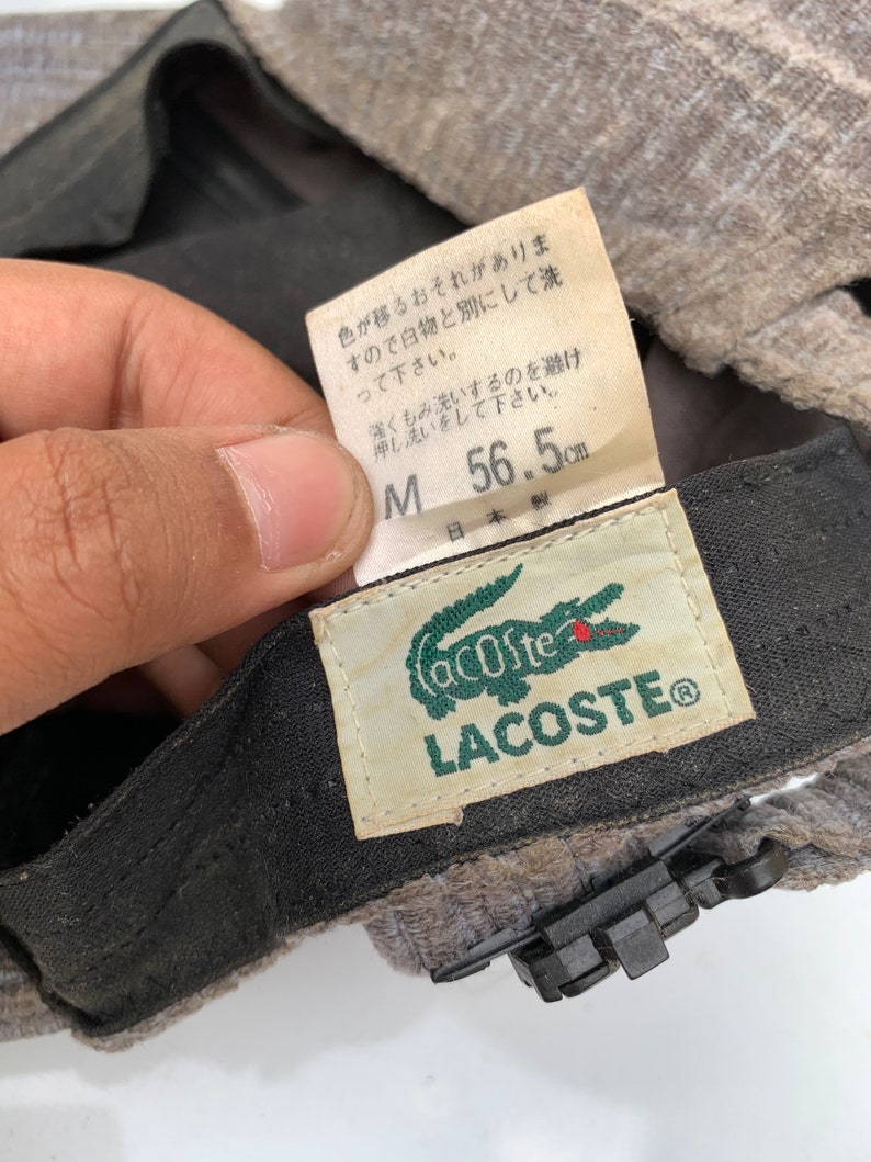 Puede incluir: Un gorro Lacoste gris y negro con un logotipo de cocodrilo verde. El gorro tiene una etiqueta de talla que dice "M 56,5 cm" y "日本製", que se traduce como "Hecho en Jap&oacute;n".
