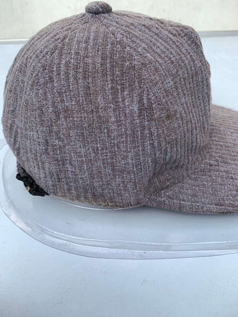Puede incluir: Una gorra de b&eacute;isbol de color marr&oacute;n con textura de costillas y un detalle de cremallera negra en el lateral.