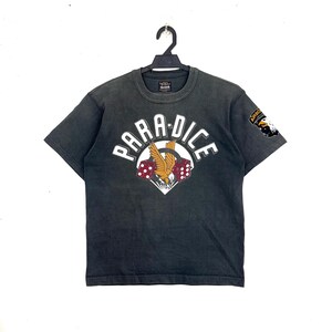 May include: Dark gray t-shirt with the word "PARADICE" in white arched text above a graphic of a bird with dice. The shirt has a distressed look and a patch on the sleeve that says "AIRBORNE".