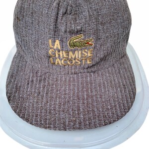 Puede incluir: Una gorra de b&eacute;isbol de pana marr&oacute;n con un logotipo de cocodrilo verde y el texto "LA CHEMISE LACOSTE" bordado en hilo dorado.