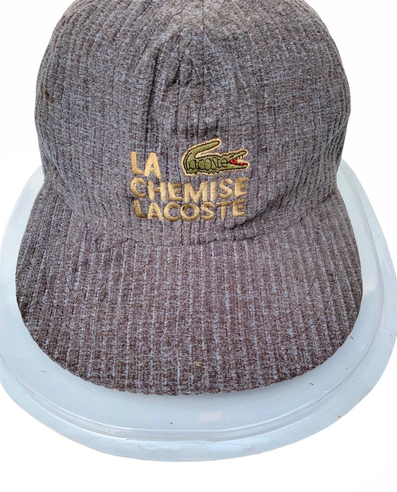 Puede incluir: Una gorra de b&eacute;isbol de pana marr&oacute;n con un logotipo de cocodrilo verde y el texto "LA CHEMISE LACOSTE" bordado en hilo dorado.