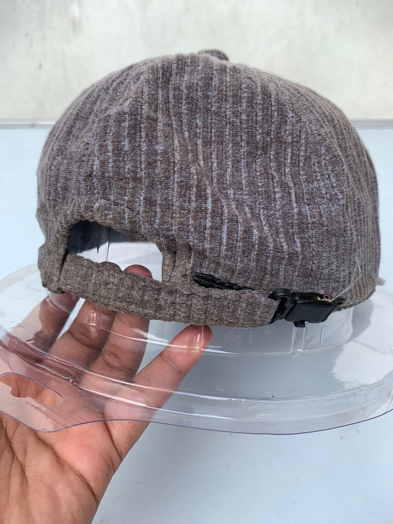Puede incluir: Una gorra de b&eacute;isbol de color marr&oacute;n y gris con textura de costillas y una correa ajustable negra.