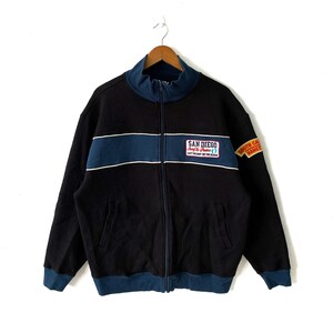 May include: Black zip-up sweatshirt with a blue and white stripe across the chest. The sweatshirt has a patch on the left chest that says "San Diego Surf & Peace" and a patch on the right sleeve that says "South Cal Hang Ten".