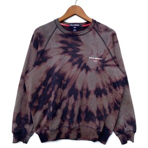 Puede incluir: Una sudadera de color marrón y negro con estampado tie-dye con el texto "POLO SPORT" y un pequeño logotipo de la bandera estadounidense en la parte delantera.