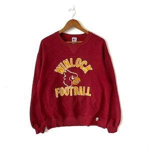 May include: Red sweatshirt with a yellow and white Winlock Football logo featuring a cardinal mascot. The sweatshirt has a crew neck and long sleeves.