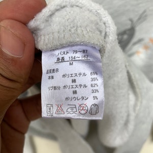 May include: A white tag with black text detailing the size and fabric composition of a gray sweatshirt. The tag reads "Bust 79-87, Height 154-162, M, Body: Polyester 65%, Cotton 35%, Rib Part: Polyester 62%, Cotton 33%, Polyurethane 5%".