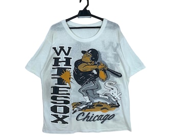 Puede incluir: Camiseta blanca con un jugador de béisbol de dibujos animados que lleva un uniforme blanco y negro y una gorra de béisbol negra. El jugador sostiene un bate de béisbol y está de pie sobre un montículo de tierra marrón. El texto "WHITE SOX Chicago" está impreso en la camiseta.