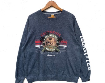 Puede incluir: Una sudadera gris de manga larga con un logotipo de Hang Ten en la manga izquierda. La parte delantera de la sudadera presenta un diseño gráfico con el texto "California" y "Hang Ten" y un diseño de palmera. El diseño gráfico también incluye el texto "Only Wild Sun Since 1962 Way of Life Fifty years of authentic surf good."