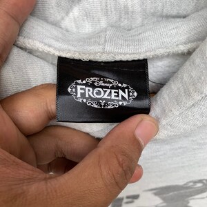 May include: A black tag with white text that reads "Disney Frozen" in a decorative font. The tag is attached to a gray hooded sweatshirt.