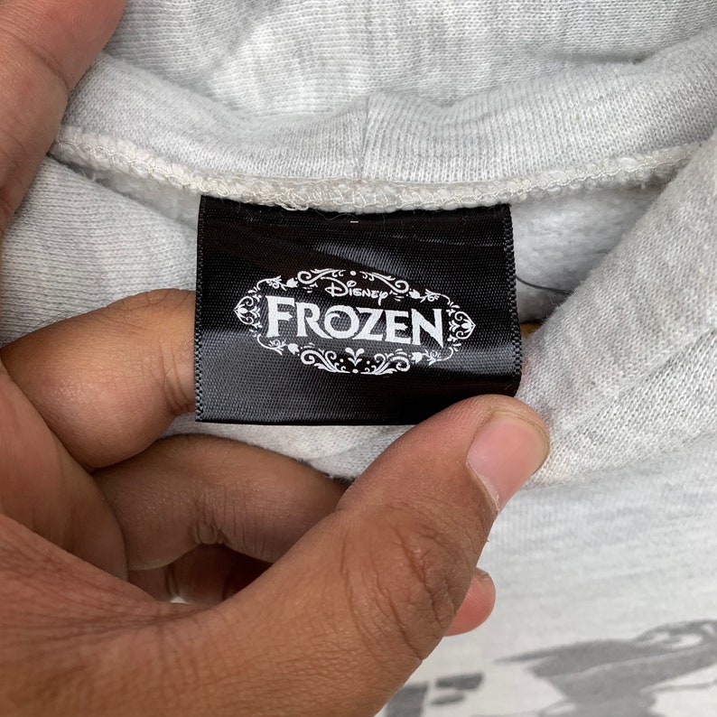 May include: A black tag with white text that reads "Disney Frozen" in a decorative font. The tag is attached to a gray hooded sweatshirt.