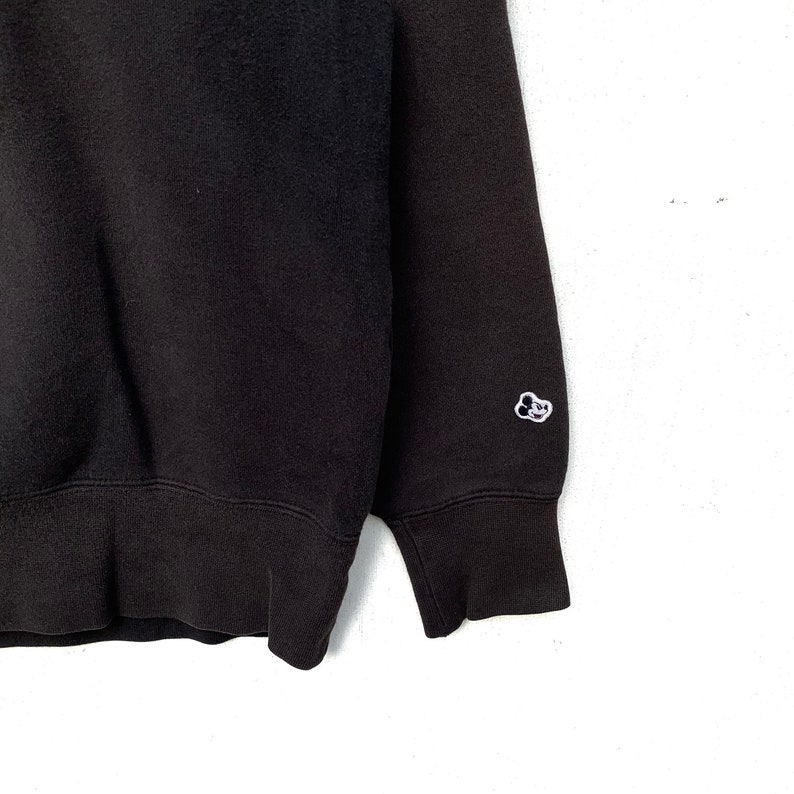 May include: Black sweatshirt with a small white embroidered logo on the left sleeve. The logo is a cartoon cloud with a face.