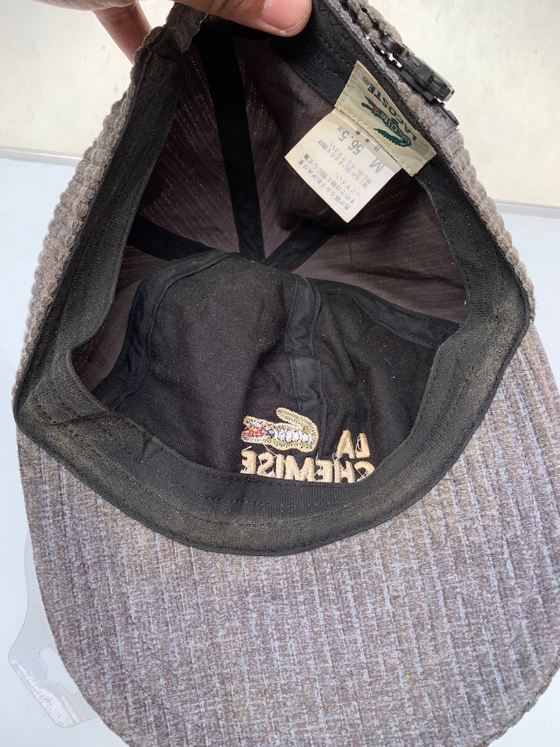 Puede incluir: Una gorra de b&eacute;isbol Lacoste gris y negra con un logotipo de cocodrilo y el texto "LA CHEMISE" en el interior. La talla es M 56,5 cm.