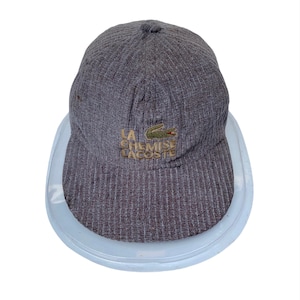 Puede incluir: Una gorra de b&eacute;isbol de color gris con textura de canal&eacute;, con un logotipo de cocodrilo verde y el texto "LA CHEMISE LACOSTE" bordado en dorado en la parte delantera.