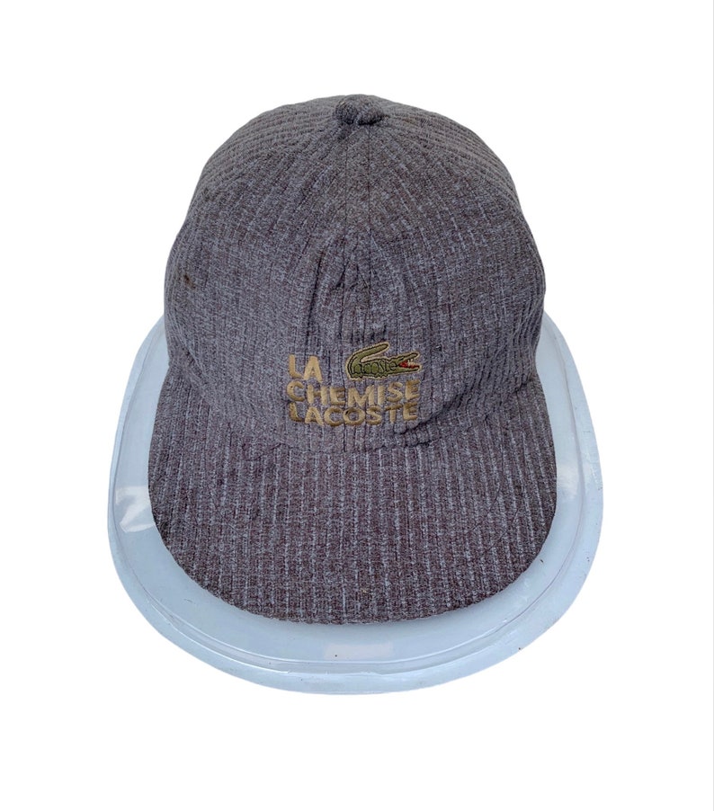 Puede incluir: Una gorra de b&eacute;isbol de color gris con textura de canal&eacute;, con un logotipo de cocodrilo verde y el texto "LA CHEMISE LACOSTE" bordado en dorado en la parte delantera.