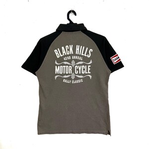 Peut inclure: Polo gris avec manches et col noirs. Le dos présente un texte blanc qui se lit "Black Hills Motor Cycle Rally Classic". Un petit écusson "Cham" est sur la manche.