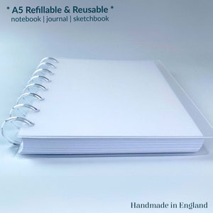 May include: A white A5 refillable and reusable notebook, journal, or sketchbook with a silver coil binding. The notebook is lying on a white surface. The text *A5 Refillable & Reusable* notebook | journal | sketchbook is printed on the cover. The text Handmade in England is printed on the bottom right corner of the image. The text sje is printed on the bottom right corner of the image.