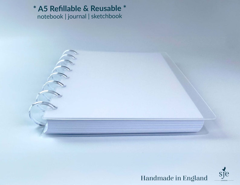 May include: A white A5 refillable and reusable notebook, journal, or sketchbook with a silver coil binding. The notebook is lying on a white surface. The text *A5 Refillable & Reusable* notebook | journal | sketchbook is printed on the cover. The text Handmade in England is printed on the bottom right corner of the image. The text sje is printed on the bottom right corner of the image.