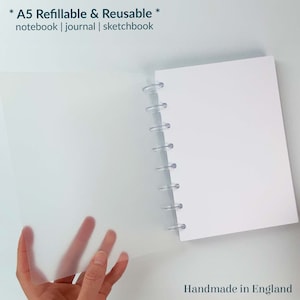 May include: A white A5 notebook with a clear plastic cover and a clear plastic spiral binding. The notebook is refillable and reusable. The text "* A5 Refillable & Reusable * notebook | journal | sketchbook" is printed on the cover. The text "Handmade in England sje" is printed on the bottom right corner of the image.