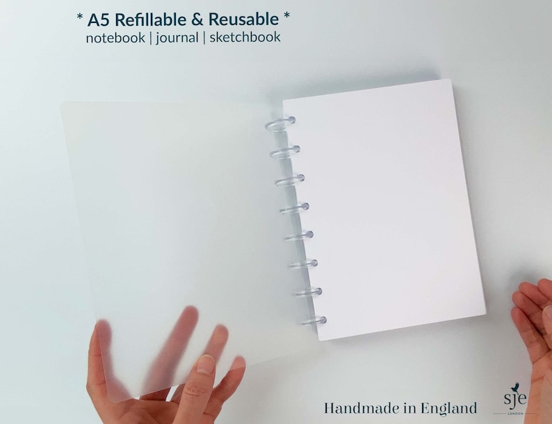 May include: A white A5 notebook with a clear plastic cover and a clear plastic spiral binding. The notebook is refillable and reusable. The text "* A5 Refillable & Reusable * notebook | journal | sketchbook" is printed on the cover. The text "Handmade in England sje" is printed on the bottom right corner of the image.