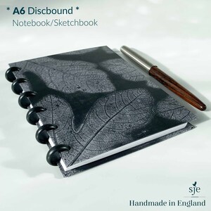 May include: A black A6 discbound notebook with a white leaf design on the cover. The notebook is closed and a silver pen is lying on top of it. The text "* A6 Discbound * Notebook/Sketchbook" is printed on the cover.