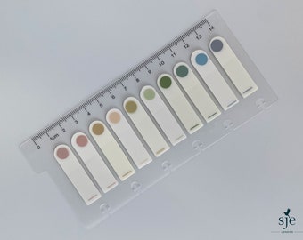 Peut inclure: Une règle en plastique transparent avec des graduations en centimètres et une série de notes autocollantes blanches. Chaque note a un cercle coloré en haut, allant du rose au bleu. La règle est en biais.