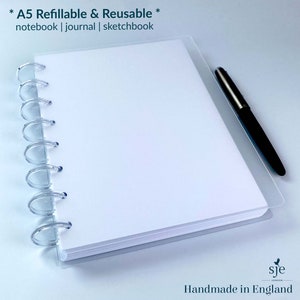 May include: A white A5 refillable and reusable notebook with a clear plastic cover and a silver coil binding. The notebook is lying flat on a white surface with a black pen next to it. The text "* A5 Refillable & Reusable * notebook | journal | sketchbook" is printed at the top of the notebook. The text "sje Handmade in England" is printed at the bottom of the notebook.