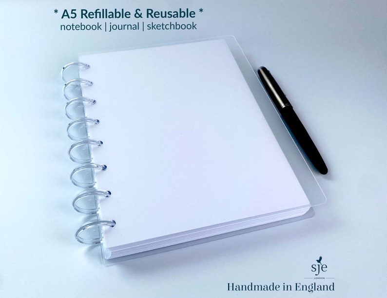 May include: A white A5 refillable and reusable notebook with a clear plastic cover and a silver coil binding. The notebook is lying flat on a white surface with a black pen next to it. The text "* A5 Refillable & Reusable * notebook | journal | sketchbook" is printed at the top of the notebook. The text "sje Handmade in England" is printed at the bottom of the notebook.