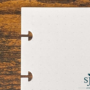 Puede incluir: Cuaderno blanco con un patr&oacute;n de cuadr&iacute;cula punteada y dos agujeros perforados en el lado izquierdo. El cuaderno tiene un logotipo en blanco y negro que dice "sje LONDON" en la esquina inferior derecha.