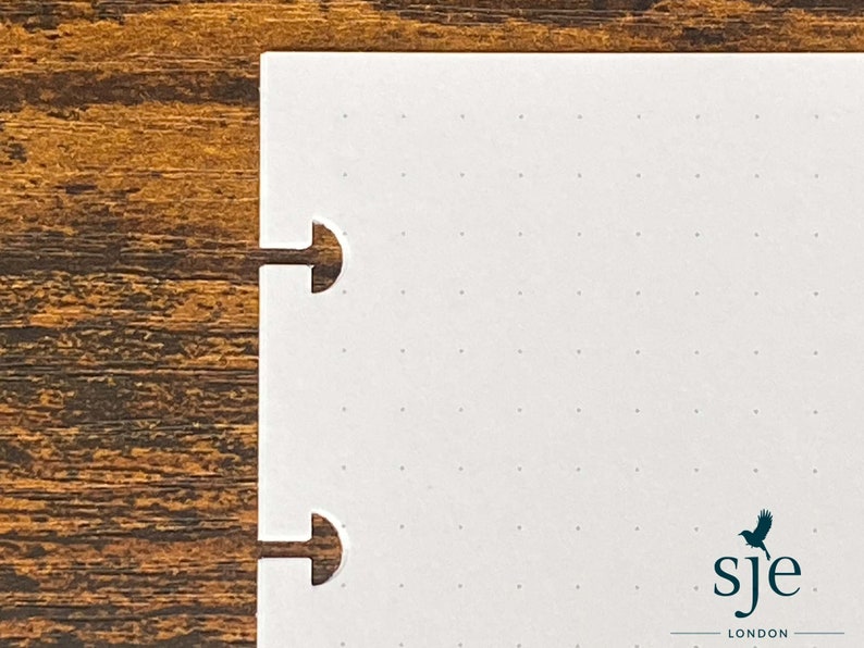 Puede incluir: Cuaderno blanco con un patr&oacute;n de cuadr&iacute;cula punteada y dos agujeros perforados en el lado izquierdo. El cuaderno tiene un logotipo en blanco y negro que dice "sje LONDON" en la esquina inferior derecha.