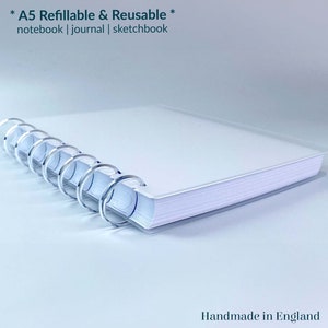May include: A white A5 refillable and reusable notebook, journal, or sketchbook with a silver coil binding. The notebook is closed and lying on a white surface. The text "* A5 Refillable & Reusable * notebook | journal | sketchbook" is printed on the cover. The text "Handmade in England" and a logo are printed on the bottom right corner of the image.