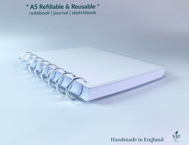 May include: A white A5 refillable and reusable notebook, journal, or sketchbook with a silver coil binding. The notebook is closed and lying on a white surface. The text "* A5 Refillable & Reusable * notebook | journal | sketchbook" is printed on the cover. The text "Handmade in England" and a logo are printed on the bottom right corner of the image.
