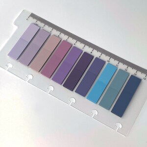 May include: A selection of sticky page markers in various shades of purple and blue, attached to a clear plastic ruler with centimetre markings. The markers are arranged in a row, each a different colour. The logo "sje LONDON" is visible in the bottom right corner.
