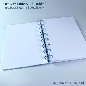 May include: An A5 refillable and reusable notebook, journal, or sketchbook with a white cover and silver binding. The text "* A5 Refillable & Reusable * notebook | journal | sketchbook" is printed on the cover. The text "Handmade in England" and a logo with the letters "sje" are printed on the bottom of the page.