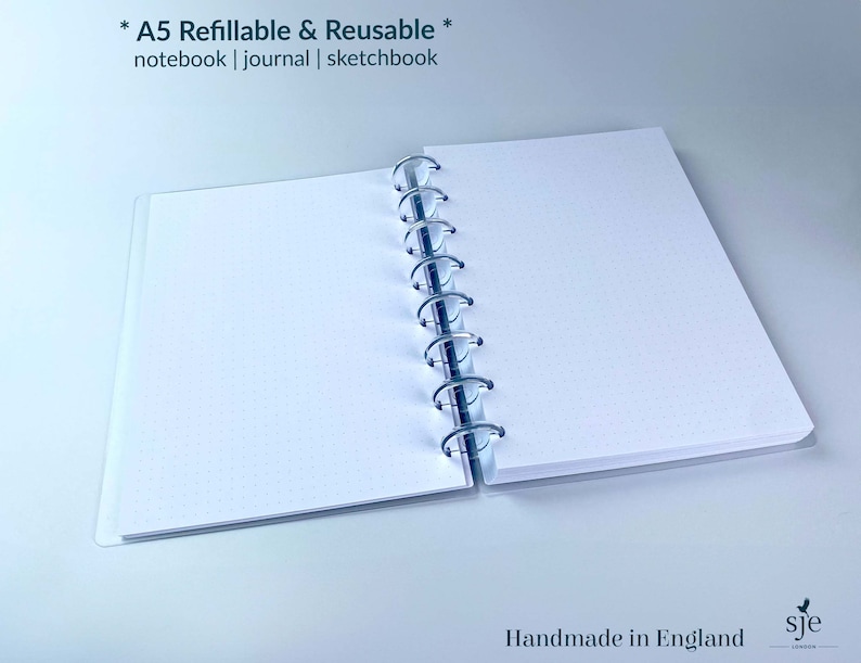 May include: An A5 refillable and reusable notebook, journal, or sketchbook with a white cover and silver binding. The text "* A5 Refillable & Reusable * notebook | journal | sketchbook" is printed on the cover. The text "Handmade in England" and a logo with the letters "sje" are printed on the bottom of the page.