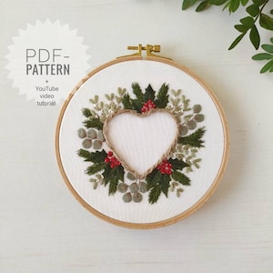 May include: Embroidered hoop art featuring a heart-shaped wreath of green leaves, red berries, and a brown twine heart. The hoop is made of wood and has a white fabric background. The text "PDF-PATTERN" and "YouTube video tutorial" is visible in the top left corner.