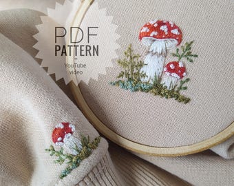 May include: A beige sweatshirt featuring embroidered red and white mushrooms on the sleeve and a wooden embroidery hoop containing a matching mushroom design. The image includes the text PDF PATTERN + YouTube video overlaid on the fabric.