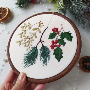 May include: A white embroidery hoop with a wooden frame, featuring a hand-stitched design of green pine branches, red berries, and green leaves.