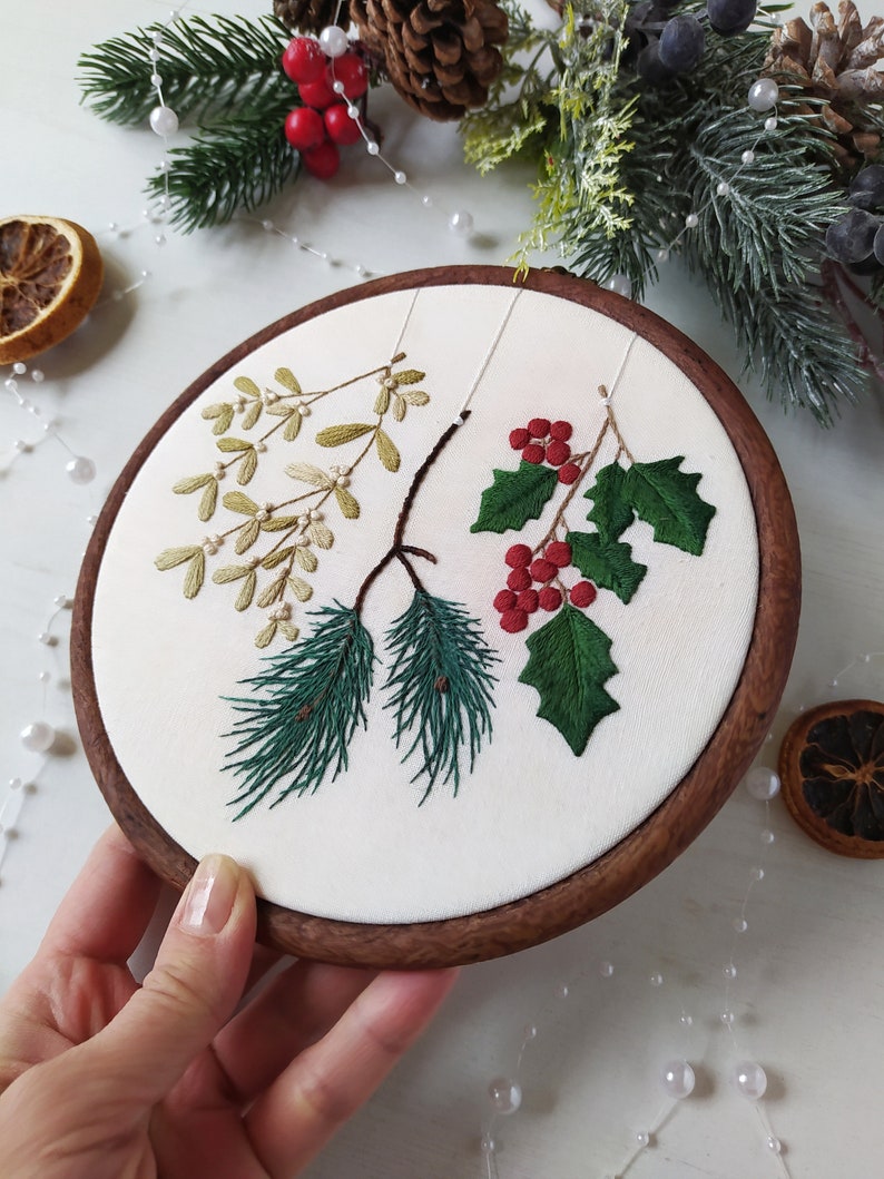 May include: A white embroidery hoop with a wooden frame, featuring a hand-stitched design of green pine branches, red berries, and green leaves.