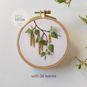 Puede incluir: Rama de abedul bordada con hojas 3D en un aro de bordado de madera. El bordado está sobre tela blanca y presenta hojas verdes y amentos amarillos. El aro está sujeto a una superficie blanca. El texto "PDF PATTERN" y "YouTube video" es visible en la imagen.