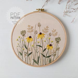 May include: Embroidered hoop art featuring a floral design with yellow flowers and green leaves on a beige background. The hoop is made of wood and has a gold metal screw.